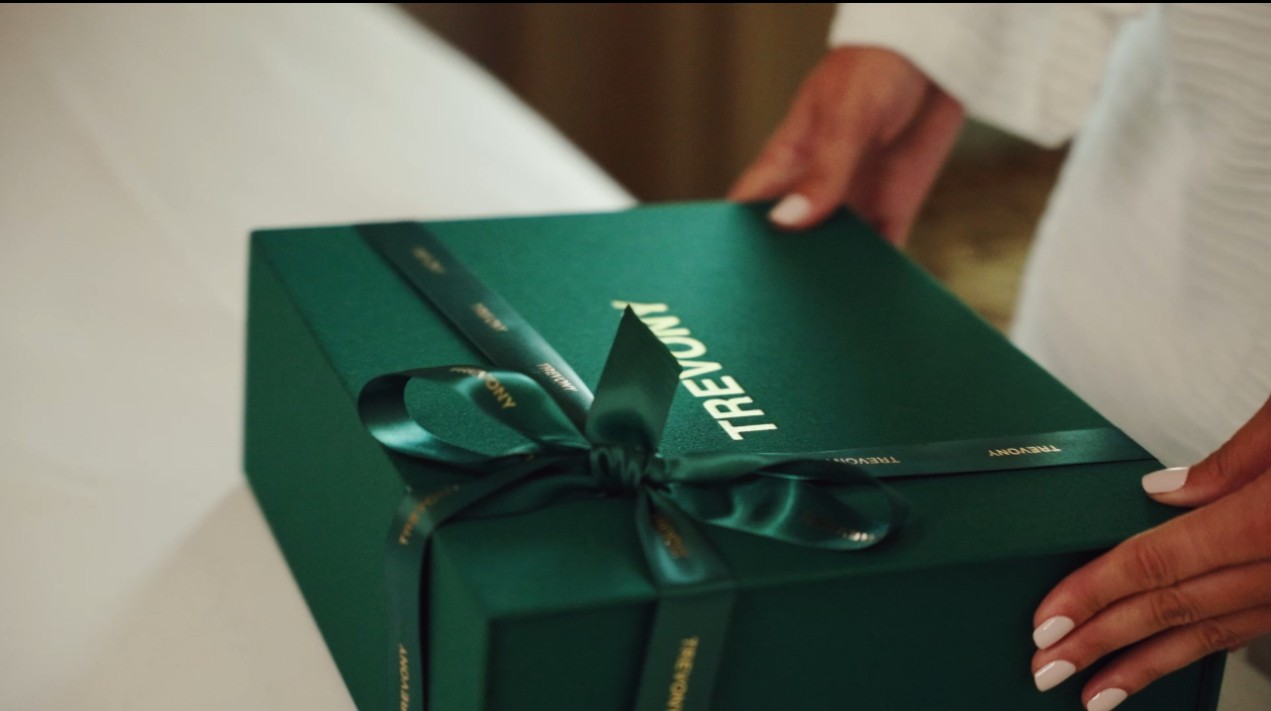 A person's hands are placing a luxurious green Trevony box with a bow on a white surface, indicating a premium unboxing experience.