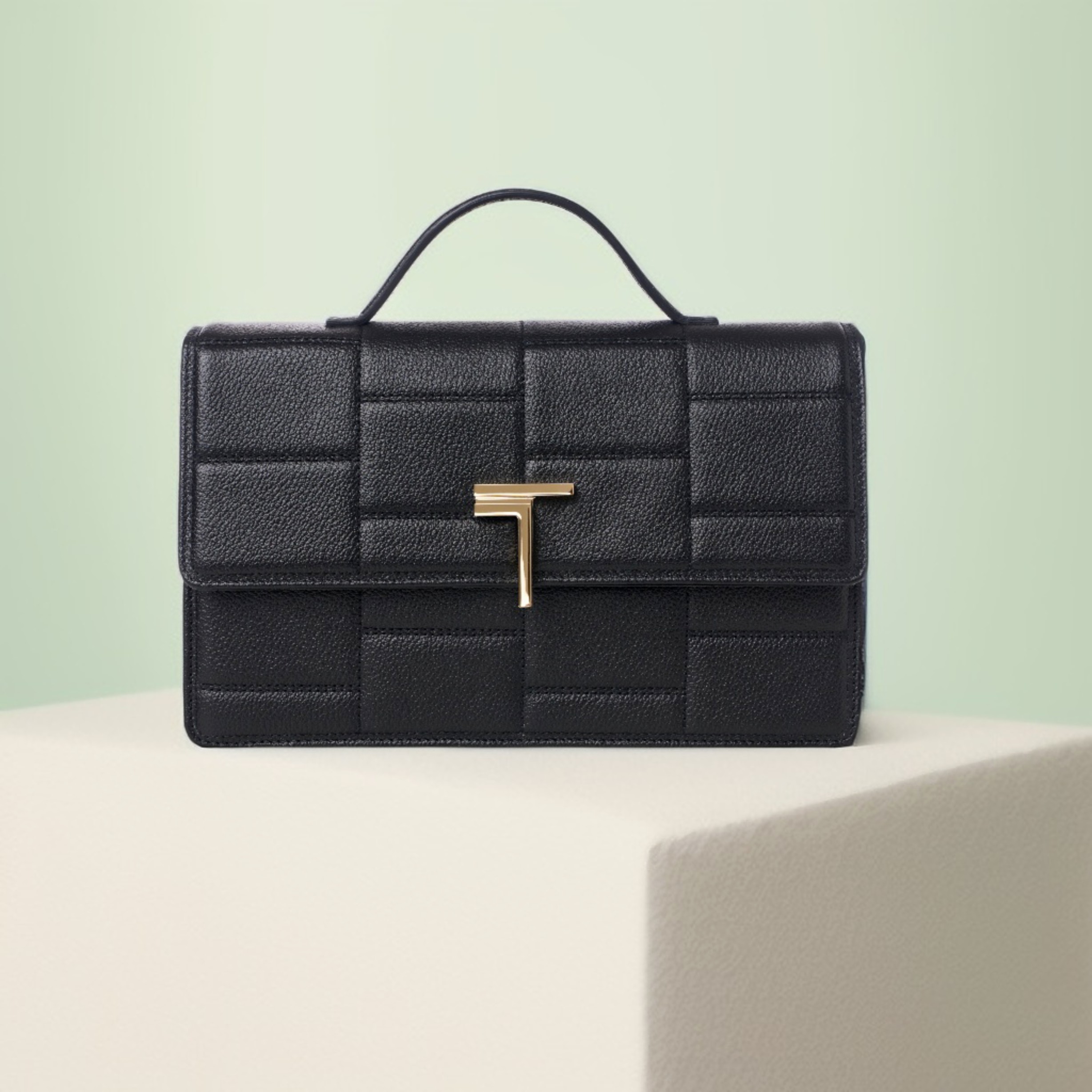 A Trevony Minerva handbag in black scratch-resistant lambskin leather with a prominent gold Trevony logo closure, displayed against a soft pastel background.